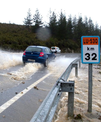Un vehículo avanza entre un torrente de agua que cruza la carretera LU 350.