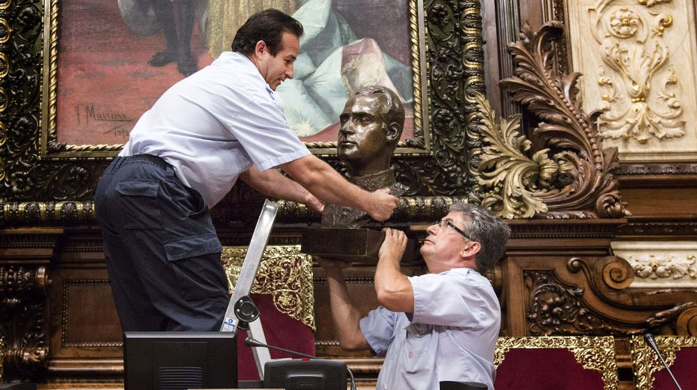Dos operarios municipales retiran el busto del Rey Juan Carlos I que hay en el Salón de la Reina Regente, donde se celebran los plenos municipales del Ayuntamiento de Barcelona.