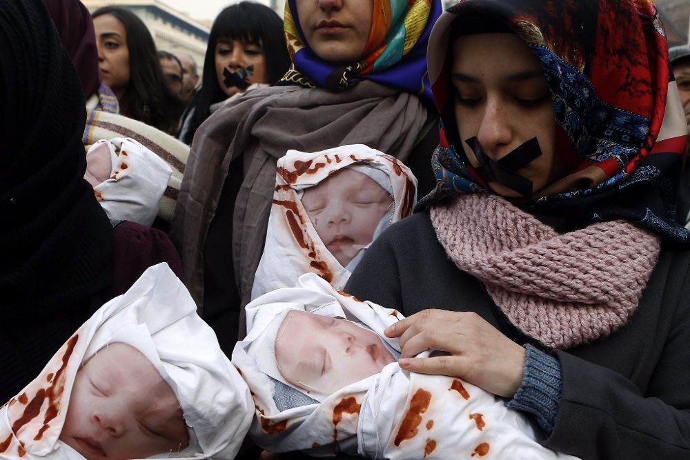 Varias personas de distintas nacionalidades llevan muñecos que representan a bebés muertos durante una marcha solidaria.