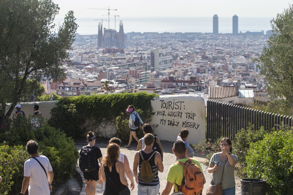 Un grupo de turistas pasea por los alrededores del Parque Guell de Barcelona ante una pancarta que dice: "Turista, tu viaje de lujo, mi miseria diaria".