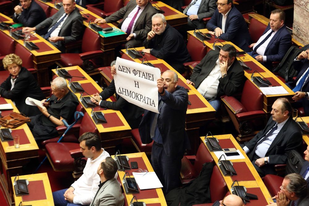 John Saridis (c), integrante del partido Unión de Centristas, sostiene un cartel en el que puede leerse ''No culpen a la Democracia'', durante la votación del Acuerdo de Prespa referente al cambio de nombre de Macedonia, este viernes en Atenas.
